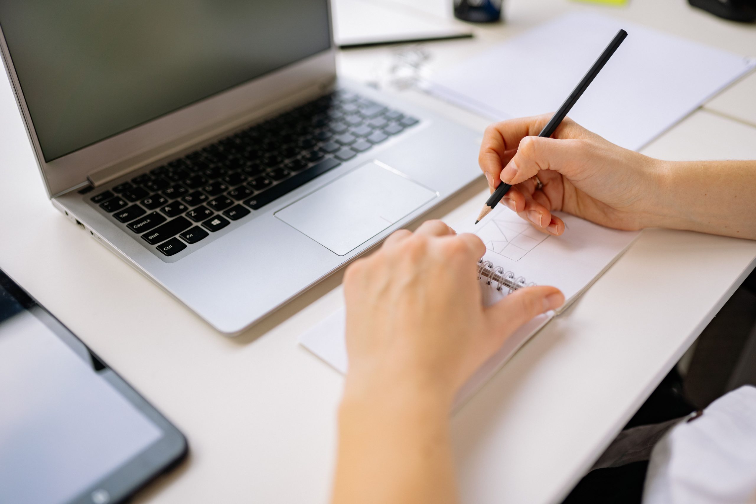 Hands sketching a website layout in a notebook next to a laptop, planning a site that converts visitors